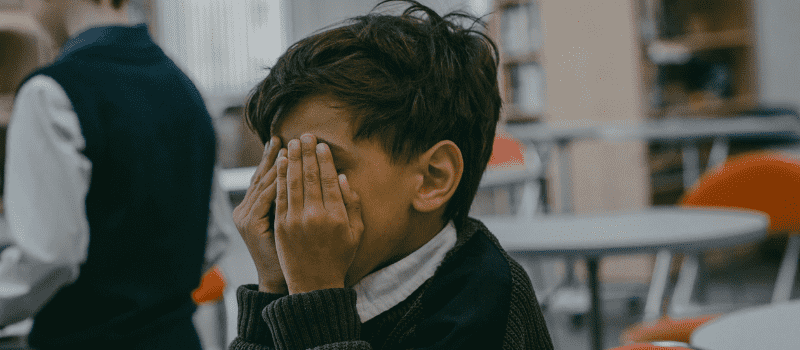 Young child sitting in a classroom covering their face with both hands, conveying distress or overwhelm